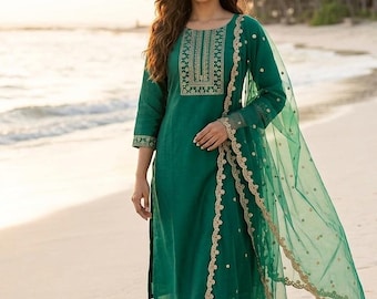 Puede incluir: Traje kurta verde esmeralda con bordados dorados en el escote y las mangas. El conjunto incluye pantalones a juego y un dupatta transparente con detalles dorados. El conjunto se usa en una playa de arena.
