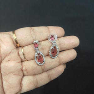 Puede incluir: Un par de pendientes colgantes plateados con una gema rosa-rojiza en forma de l&aacute;grima, rodeada de peque&ntilde;os cristales transparentes. Cierre de tuerca. Se muestran sobre un fondo oscuro.