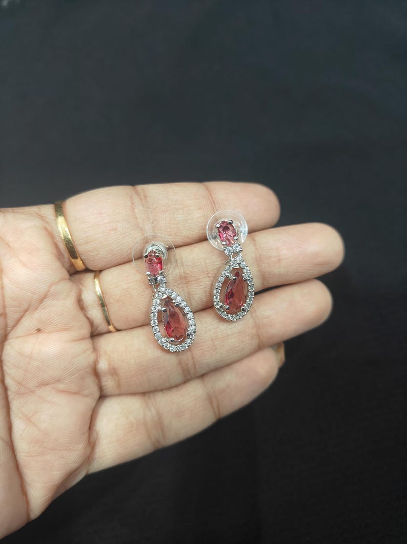 Puede incluir: Un par de pendientes colgantes plateados con una gema rosa-rojiza en forma de l&aacute;grima, rodeada de peque&ntilde;os cristales transparentes. Cierre de tuerca. Se muestran sobre un fondo oscuro.