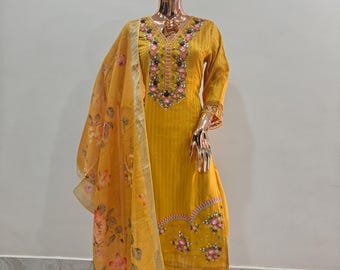 Puede incluir: Conjunto salwar kameez amarillo dorado sobre un maniquí. El atuendo presenta intrincados bordados florales en el escote, las mangas y los pantalones. Un dupatta transparente a juego con un estampado floral cae sobre el hombro del maniquí.