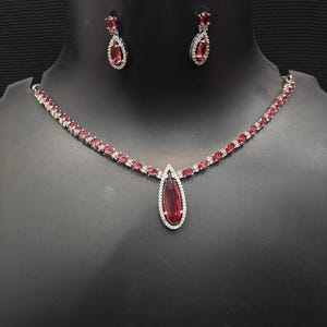 Puede incluir: Un conjunto de collar y pendientes plateados. El collar presenta un colgante en forma de l&aacute;grima con una gran piedra preciosa de color rojo intenso, rodeada de peque&ntilde;as piedras transparentes. Los pendientes a juego tienen un dise&ntilde;o similar. El conjunto se exhibe sobre un maniqu&iacute; negro.