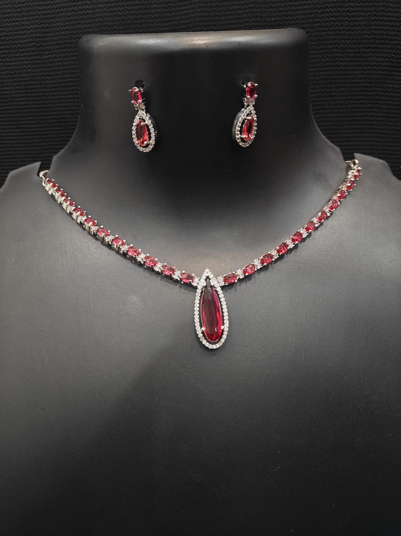 Puede incluir: Un conjunto de collar y pendientes plateados. El collar presenta un colgante en forma de l&aacute;grima con una gran piedra preciosa de color rojo intenso, rodeada de peque&ntilde;as piedras transparentes. Los pendientes a juego tienen un dise&ntilde;o similar. El conjunto se exhibe sobre un maniqu&iacute; negro.