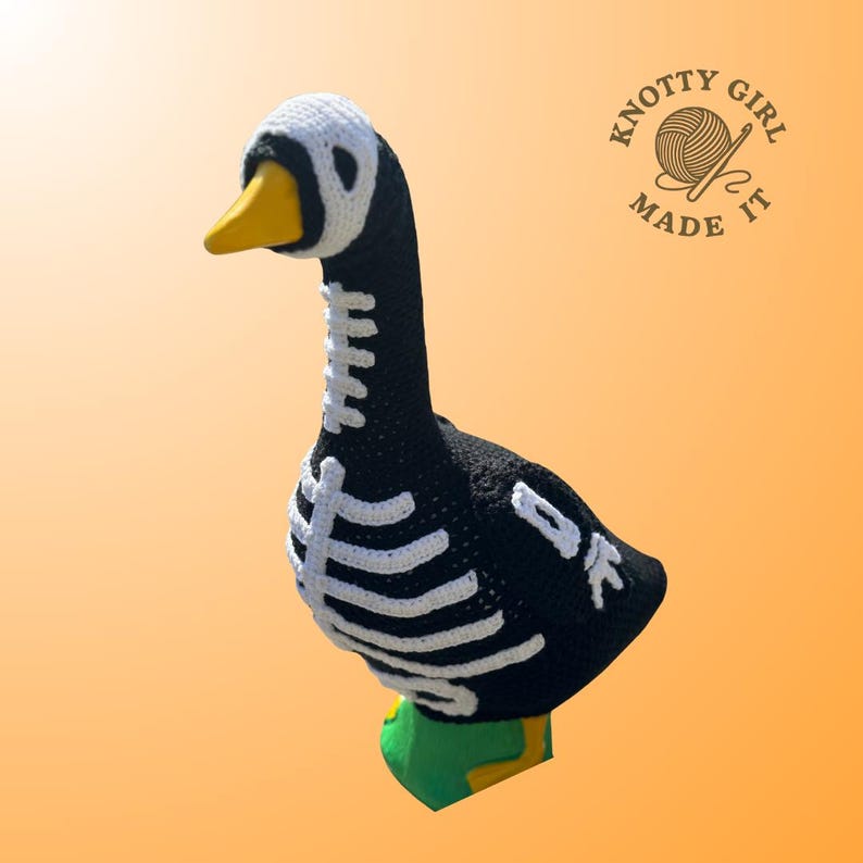 K&ouml;nnte beinhalten: Handgeh&auml;kelte Gans in Schwarz mit wei&szlig;en Skelett-Details. Die Gans hat einen gelben Schnabel und eine wei&szlig;e Kopfbedeckung. Der Sockel ist gr&uuml;n und gelb. Das Logo "KNOTTY GIRL MADE IT" ist zu sehen.