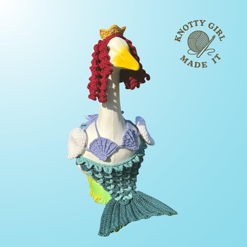 K&ouml;nnte beinhalten: Eine skurrile Gans, die sich als Meerjungfrau verkleidet hat, mit einem geh&auml;kelten Schwanz, einem Muschel-Bikini und roten Locken. Die Gans tr&auml;gt eine goldene Krone. Das Logo "Knotty Girl Made It" ist sichtbar.