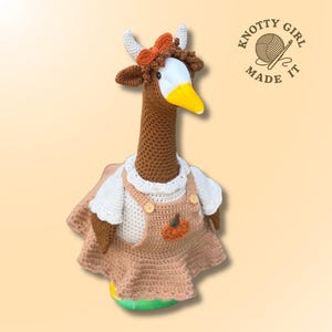 May include: A brown crocheted goose doll with a white face and yellow beak, wearing a white shirt and tan overalls with a small orange pumpkin appliqué. The goose has small white horns and a brown bow on its head. Text reads KNOTTY GIRL MADE IT.