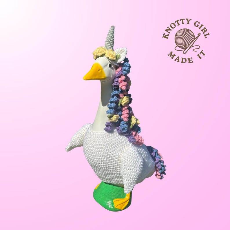 May include: A white goose figurine with a yellow beak and feet, adorned with a crocheted unicorn horn, a floral crown, and a colorful mane. The goose is wearing a green base with yellow feet. The text "Knotty Girl Made It" is visible.