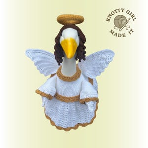 May include: A goose figurine dressed as an angel, wearing a white crochet dress with gold trim, white wings, and a brown halo. The goose has brown hair and a yellow beak. The text "KNOTTY GIRL MADE IT" is in the upper right corner.