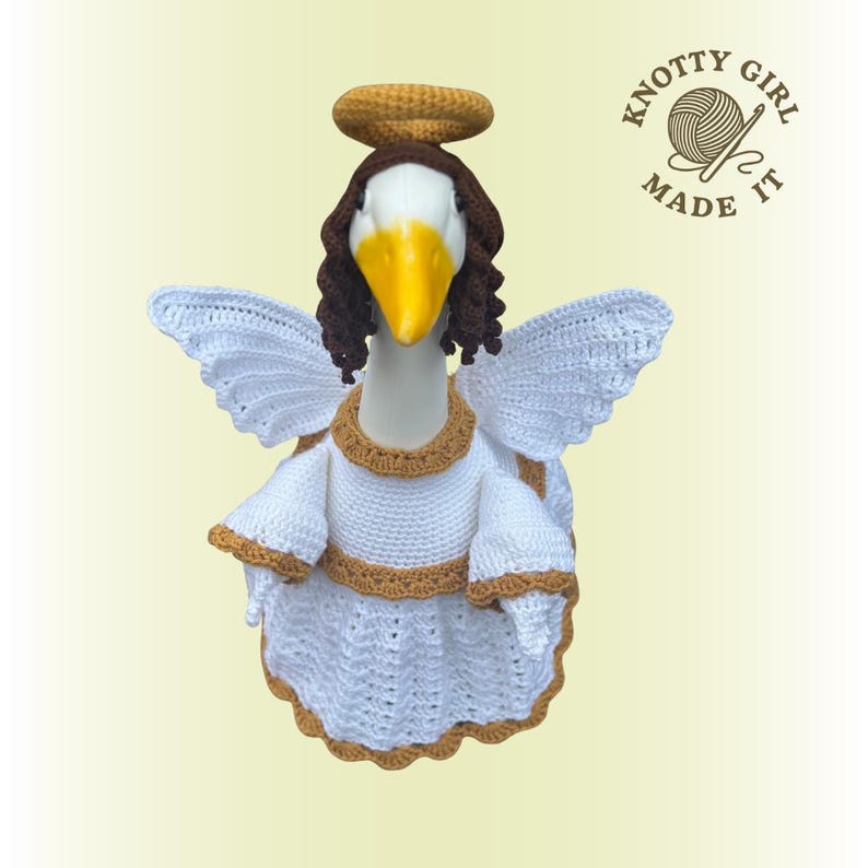 May include: A goose figurine dressed as an angel, wearing a white crochet dress with gold trim, white wings, and a brown halo. The goose has brown hair and a yellow beak. The text "KNOTTY GIRL MADE IT" is in the upper right corner.