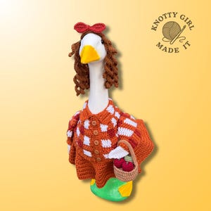 May include: A white goose figurine wearing a crocheted outfit consisting of a red and white checkered shirt with brown buttons, a solid orange skirt, and a matching red bow headband with brown curly hair. It holds a small tan basket with red apples.