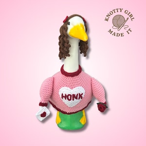 Könnte beinhalten: Eine handgefertigte Häkelgans mit einem rosa Pullover mit einem weißen Herz mit der Aufschrift "HONK". Die Gans hat braune Locken, eine rote Schleife und einen gelben Schnabel. Sie hält einen kleinen weißen Umschlag mit einem roten Herzen.