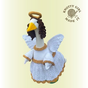 May include: A handmade crochet goose dressed as an angel, featuring white wings, a halo, and a white dress with gold trim. The goose has a yellow beak and brown hair. The text "KNOTTY GIRL MADE IT" is visible.