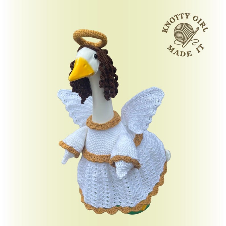 May include: A handmade crochet goose dressed as an angel, featuring white wings, a halo, and a white dress with gold trim. The goose has a yellow beak and brown hair. The text "KNOTTY GIRL MADE IT" is visible.