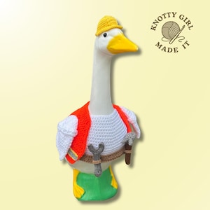 May include: A goose figurine dressed in a handmade crochet outfit. The goose wears a yellow hat, an orange vest, and a white crochet shirt. It has a tool belt with a wrench and hammer. The logo "Knotty Girl Made It" is in the upper right corner.