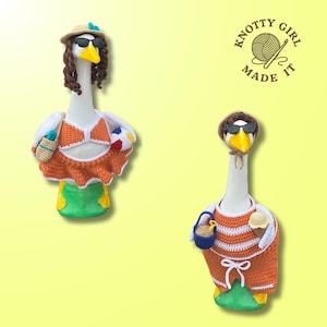 May include: Two goose figurines, each adorned in crochet beach attire. One wears an orange bikini, skirt, and sunglasses, holding a beach ball and bag. The other sports an orange and white striped top, shorts, and sunglasses, carrying a bucket and ice cream cone.