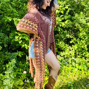 May include: A long, open-front crochet shawl in shades of brown, yellow, and pink. The shawl is worn over a brown t-shirt and denim shorts. The person is wearing brown cowboy boots and standing in a grassy area.