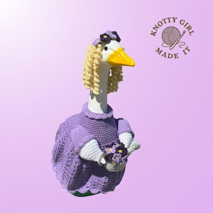 May include: A whimsical goose figurine wearing a crocheted purple dress, a matching hat, and a blonde wig. The goose holds a basket of purple flowers. The logo "Knotty Girl Made It" is in the upper right corner.