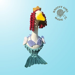 Può includere: Una scultura di oca stravagante vestita da sirena, con coda all'uncinetto, reggiseno a conchiglia e capelli ricci rossi. L'oca indossa una corona d'oro e ha un becco giallo. La scultura è ambientata su uno sfondo azzurro chiaro, con il testo "KNOTTY GIRL MADE IT".