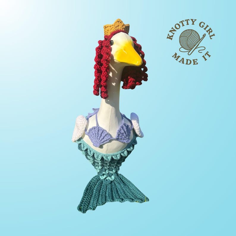 K&ouml;nnte beinhalten: Eine skurrile G&auml;nse-Skulptur, die als Meerjungfrau gekleidet ist, mit einem geh&auml;kelten Schwanz, einem Muschel-BH und roten Locken. Die Gans tr&auml;gt eine goldene Krone und hat einen gelben Schnabel. Die Skulptur steht vor einem hellblauen Hintergrund, mit dem Text "KNOTTY GIRL MADE IT".