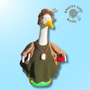 May include: A white goose figurine dressed in a crocheted outfit, including a green sweater, tan cardigan, and a brown wig with a pencil. The goose wears glasses and a pendant. It holds a red apple and is on a green and yellow base.