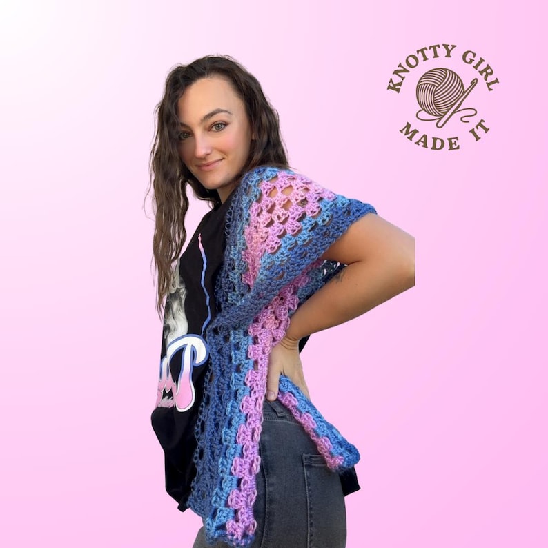 Pode incluir: Um xale de croch&ecirc; feito &agrave; m&atilde;o em tons de azul, rosa e roxo. O design de tecido aberto &eacute; vis&iacute;vel, e o xale &eacute; drapeado sobre os ombros. A imagem inclui o texto "KNOTTY GIRL MADE IT" em um logotipo circular.
