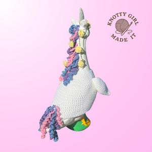 May include: A white crocheted goose figurine transformed into a unicorn, featuring a horn, mane, and tail in pastel colors. The goose wears a crocheted sweater and stands on a green base with yellow accents. The logo "KNOTTY GIRL MADE IT" is in the upper right corner.