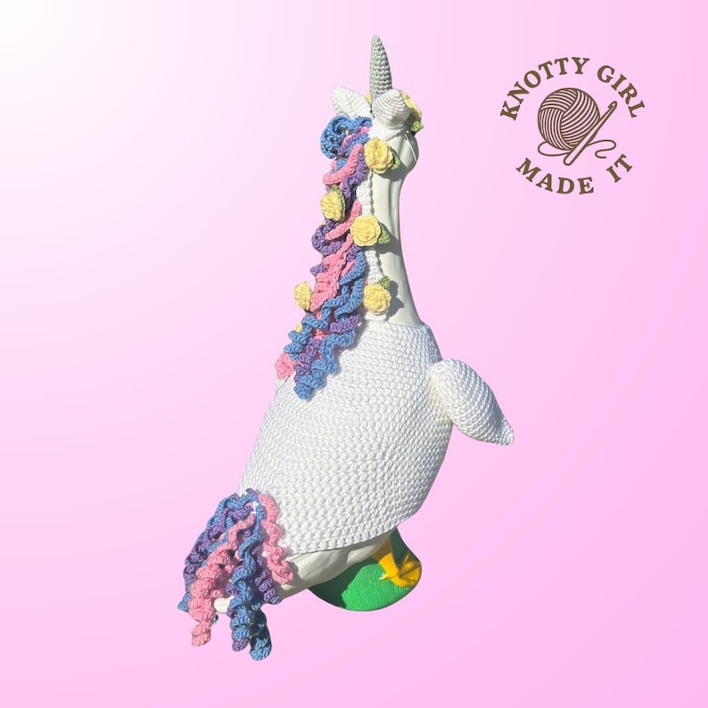 May include: A white crocheted goose figurine transformed into a unicorn, featuring a horn, mane, and tail in pastel colors. The goose wears a crocheted sweater and stands on a green base with yellow accents. The logo "KNOTTY GIRL MADE IT" is in the upper right corner.