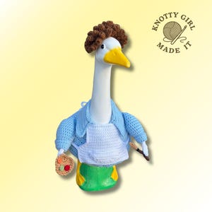 May include: A white goose figurine with a yellow beak, wearing a crocheted blue jacket, white apron, and green pants. It has a brown crocheted wig and holds a small palette and paintbrush. The text "KNOTTY GIRL MADE IT" is in the upper right corner.