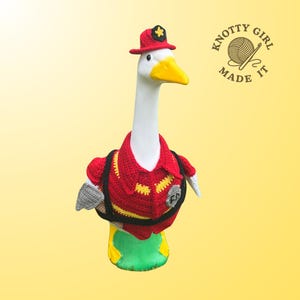 May include: A white goose figurine wearing a handmade red firefighter uniform, complete with a hat and a black backpack. The uniform has yellow and black accents. The goose has a yellow beak and is set against a yellow background.