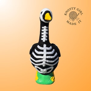 K&ouml;nnte beinhalten: Handgefertigte H&auml;kelgans-Figur in Schwarz mit wei&szlig;em Skelett-Design. Die Gans hat einen wei&szlig;en Kopf, einen gelben Schnabel und steht auf einer gr&uuml;nen Basis mit gelben F&uuml;&szlig;en. Das Logo "Knotty Girl Made It" ist sichtbar. Halloween-Dekoration.