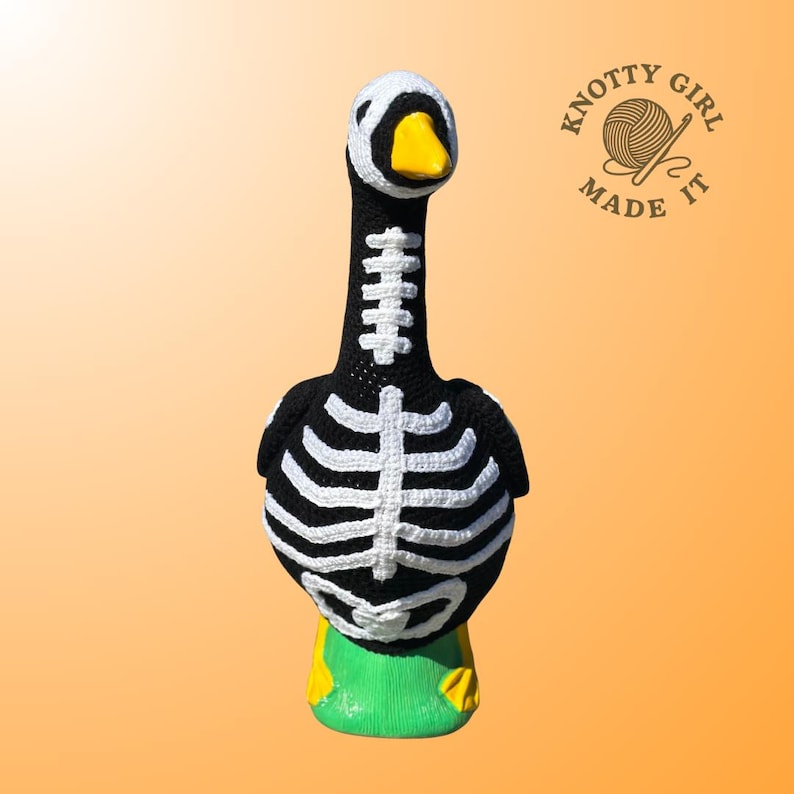 K&ouml;nnte beinhalten: Handgefertigte H&auml;kelgans-Figur in Schwarz mit wei&szlig;em Skelett-Design. Die Gans hat einen wei&szlig;en Kopf, einen gelben Schnabel und steht auf einer gr&uuml;nen Basis mit gelben F&uuml;&szlig;en. Das Logo "Knotty Girl Made It" ist sichtbar. Halloween-Dekoration.