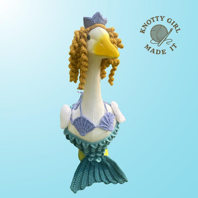K&ouml;nnte beinhalten: Eine skurrile G&auml;nsefigur als Meerjungfrau verkleidet. Die Gans tr&auml;gt einen geh&auml;kelten Muschel-Bikini, eine blaue Krone und lockiges gelbes Haar. Die Meerjungfrauenschwanzflosse ist t&uuml;rkis. Das Logo "Knotty Girl Made It" ist zu sehen.