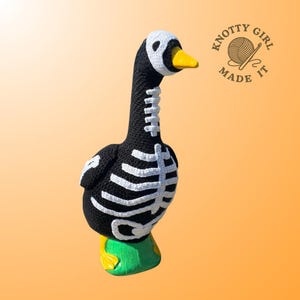 K&ouml;nnte beinhalten: Eine handgeh&auml;kelte Gans in Schwarz mit wei&szlig;en Skelett-Details. Die Gans hat einen gelben Schnabel, einen wei&szlig;en Kopf und gr&uuml;ne und gelbe F&uuml;&szlig;e. Das Logo "Knotty Girl Made It" befindet sich oben rechts.