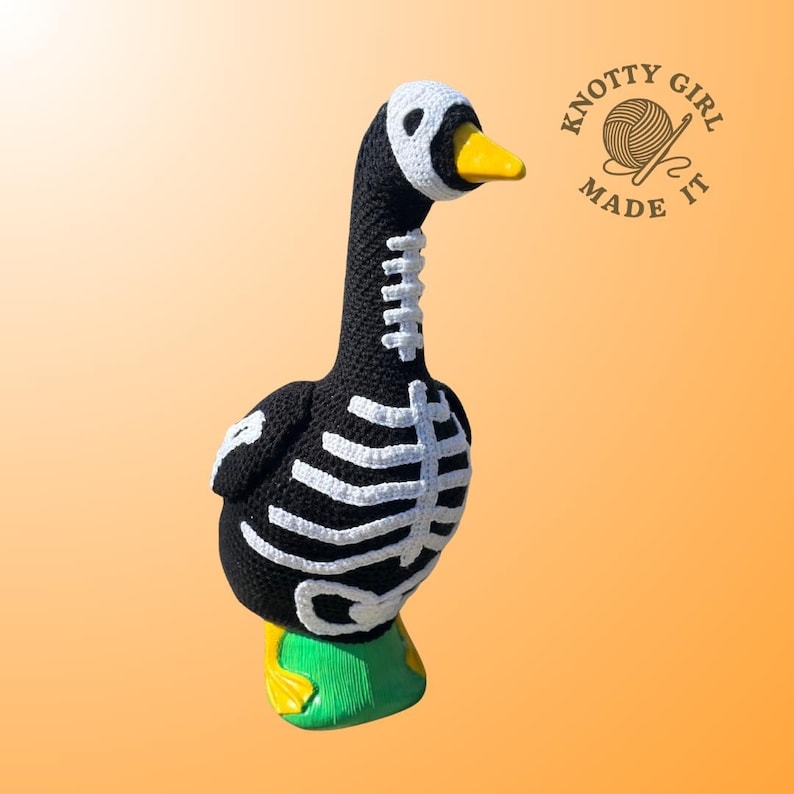 K&ouml;nnte beinhalten: Eine handgeh&auml;kelte Gans in Schwarz mit wei&szlig;en Skelett-Details. Die Gans hat einen gelben Schnabel, einen wei&szlig;en Kopf und gr&uuml;ne und gelbe F&uuml;&szlig;e. Das Logo "Knotty Girl Made It" befindet sich oben rechts.