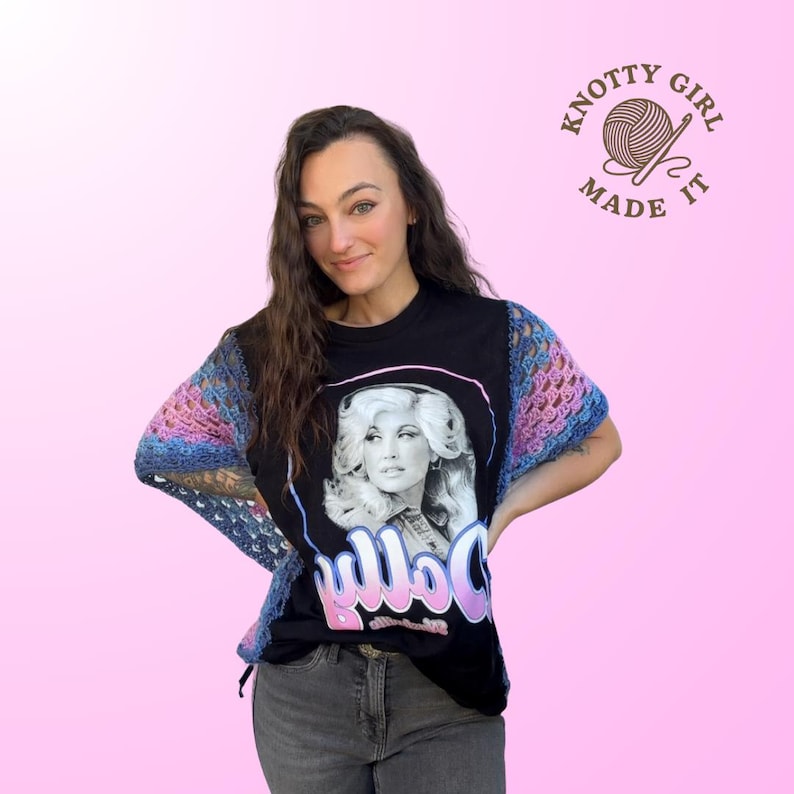 Pode incluir: Camiseta preta com um retrato de Dolly Parton e o nome "Dolly" em escrita rosa e branca. A camisa tem mangas de croch&ecirc; em tons de azul, rosa e roxo. A imagem tamb&eacute;m inclui o texto "Knotty Girl Made It".