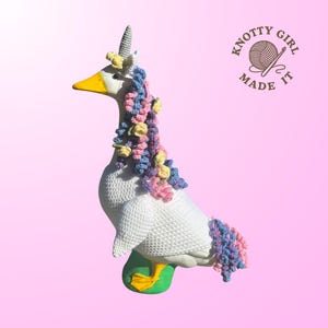 May include: A whimsical white goose figurine, adorned with a crocheted mane and tail in pastel colors, and a unicorn horn. The goose has a yellow beak and feet, and is standing on a green base. The logo "Knotty Girl Made It" is visible.
