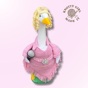 May include: A white goose figurine wearing a crocheted light pink poncho with fringe, a matching pink collar, and a belt featuring a large rhinestone buckle. The goose has blonde curly yarn hair, a yellow beak, and holds a small grey microphone. Text reads KNOTTY GIRL MADE IT.