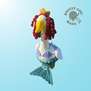 K&ouml;nnte beinhalten: Eine skurrile G&auml;nsefigur, die als Meerjungfrau verkleidet ist, komplett mit geh&auml;keltem t&uuml;rkisfarbenem Schwanz, Muschelbikini und roten Locken. Eine goldene Krone sitzt auf ihrem Kopf. Das Logo "Knotty Girl Made It" ist zu sehen.