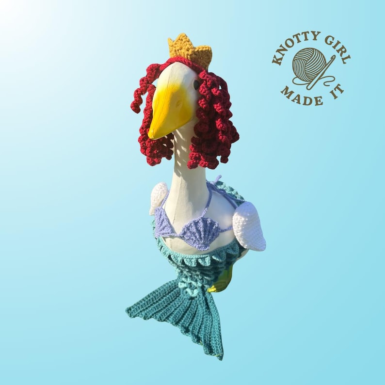 K&ouml;nnte beinhalten: Eine skurrile G&auml;nsefigur, die als Meerjungfrau verkleidet ist, komplett mit geh&auml;keltem t&uuml;rkisfarbenem Schwanz, Muschelbikini und roten Locken. Eine goldene Krone sitzt auf ihrem Kopf. Das Logo "Knotty Girl Made It" ist zu sehen.