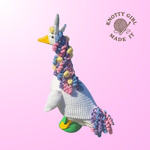 May include: A white goose figurine, crocheted to resemble a unicorn, with a yellow beak and a horn. The goose is adorned with a colorful mane and tail of crocheted flowers in shades of pink, blue, and yellow. The text "KNOTTY GIRL MADE IT" is in the upper right corner.
