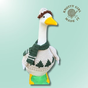 May include: A white goose figurine wearing a beige crocheted sweater with dark green pine tree patterns, a matching dark green scarf with white stripes, and a small beige hat with a brown brim. The text Knotty Girl Made It is visible.