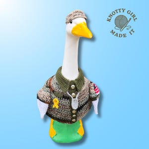 May include: A white goose figurine wearing a crocheted camouflage shirt with a dark green collar, silver buttons, a yellow ribbon pin, and a small American flag patch on the sleeve. The goose wears a matching tan crocheted hat and a small gray tag.