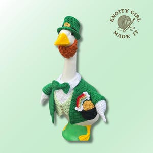 May include: A crocheted goose in a leprechaun outfit, featuring a green hat, jacket, and bow tie. The goose has a red beard and a pot of gold with a rainbow. The logo "Knotty Girl Made It" is visible in the top right corner.