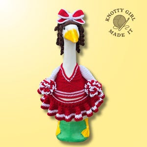 May include: A handmade crochet goose wearing a red and white cheerleader outfit, complete with pom-poms and a bow. The goose has brown curly hair and a yellow beak. The goose is standing on a green base.