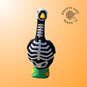 K&ouml;nnte beinhalten: Eine geh&auml;kelte Gans in Schwarz mit wei&szlig;en Skelett-Details. Die Gans hat einen wei&szlig;en Kopf, einen gelben Schnabel und eine gr&uuml;n-gelbe Basis. Der Text "KNOTTY GIRL MADE IT" befindet sich in der oberen rechten Ecke.