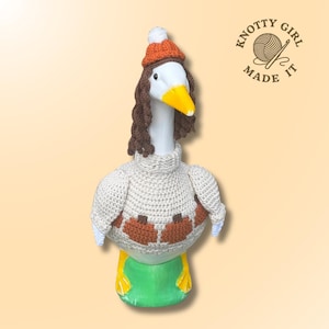 May include: A white goose figurine wearing a beige crocheted jumper with orange pumpkin motifs, a brown crocheted wig with long curls, and an orange beanie with a white pom-pom. The text Knotty Girl Made It is visible in the upper right corner.