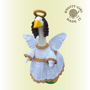 May include: A white goose figurine dressed as an angel, complete with crocheted wings, a halo, and a dress with gold trim. The goose has brown hair and a yellow beak. The text "KNOTTY GIRL MADE IT" is in the upper right corner.