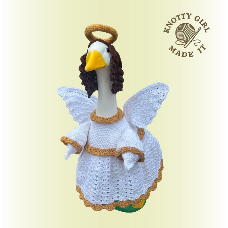 May include: A white goose figurine dressed as an angel, complete with crocheted wings, a halo, and a dress with gold trim. The goose has brown hair and a yellow beak. The text "KNOTTY GIRL MADE IT" is in the upper right corner.