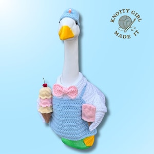 May include: A white goose figurine wearing a light blue crocheted hat, a white shirt, a light blue apron, and a pink bow tie. The goose holds a crocheted ice cream cone with a red cherry and a pink rectangular treat. Text reads KNOTTY GIRL MADE IT.