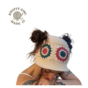 May include: Cream-colored crochet hat with floral designs in teal, pink, and cream. The hat features the text "KNOTTY GIRL MADE IT" and is worn on the head. The hat is a unique, handmade accessory.