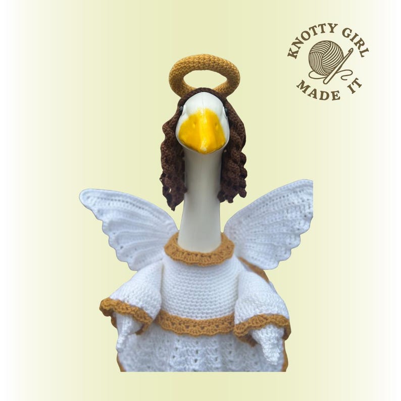May include: A handcrafted goose figurine dressed as an angel, featuring white crochet wings, a white dress with gold trim, and a brown halo and hair. The goose has a yellow beak and the text "KNOTTY GIRL MADE IT" is visible.